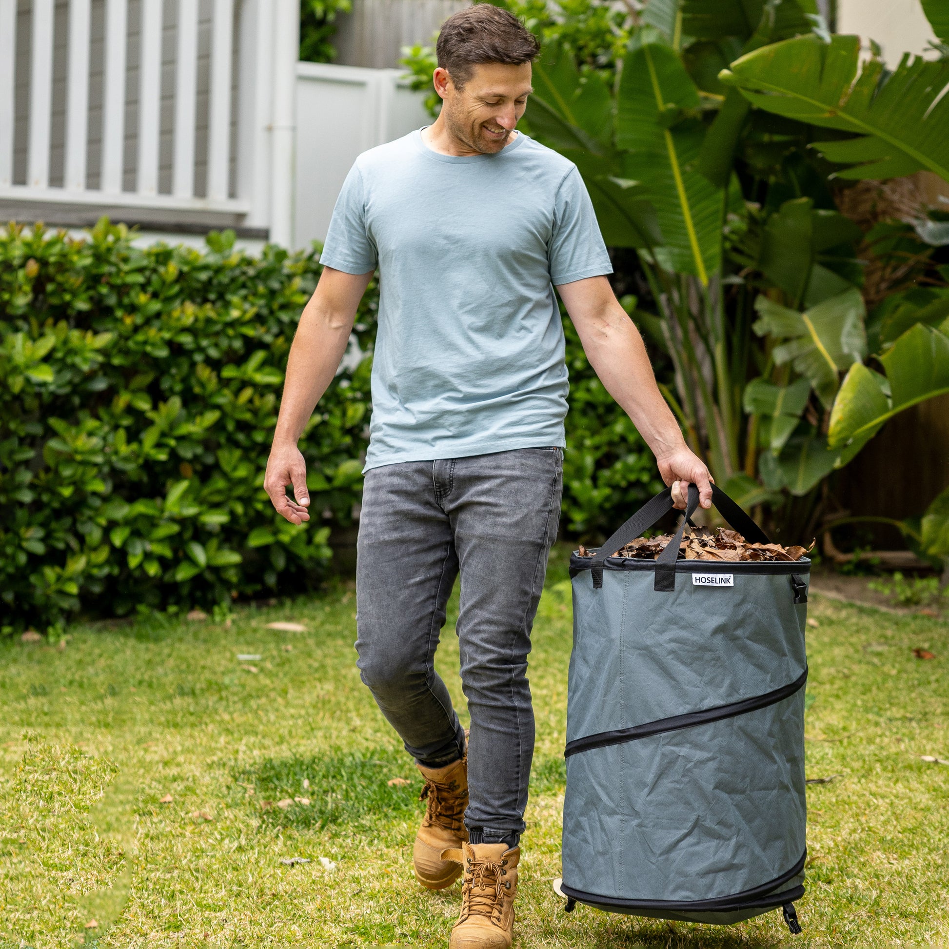 Person in blue shirt carrying charcoal colured garden waster bag full of brown leaves over a green lawn