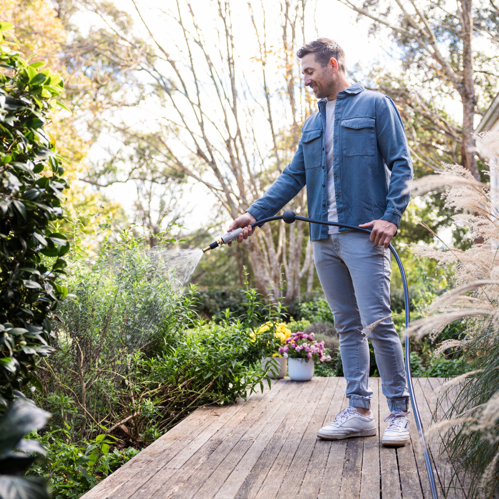 person standing on deck watering green bushes with the brass garden hose nozzle with trees in the background