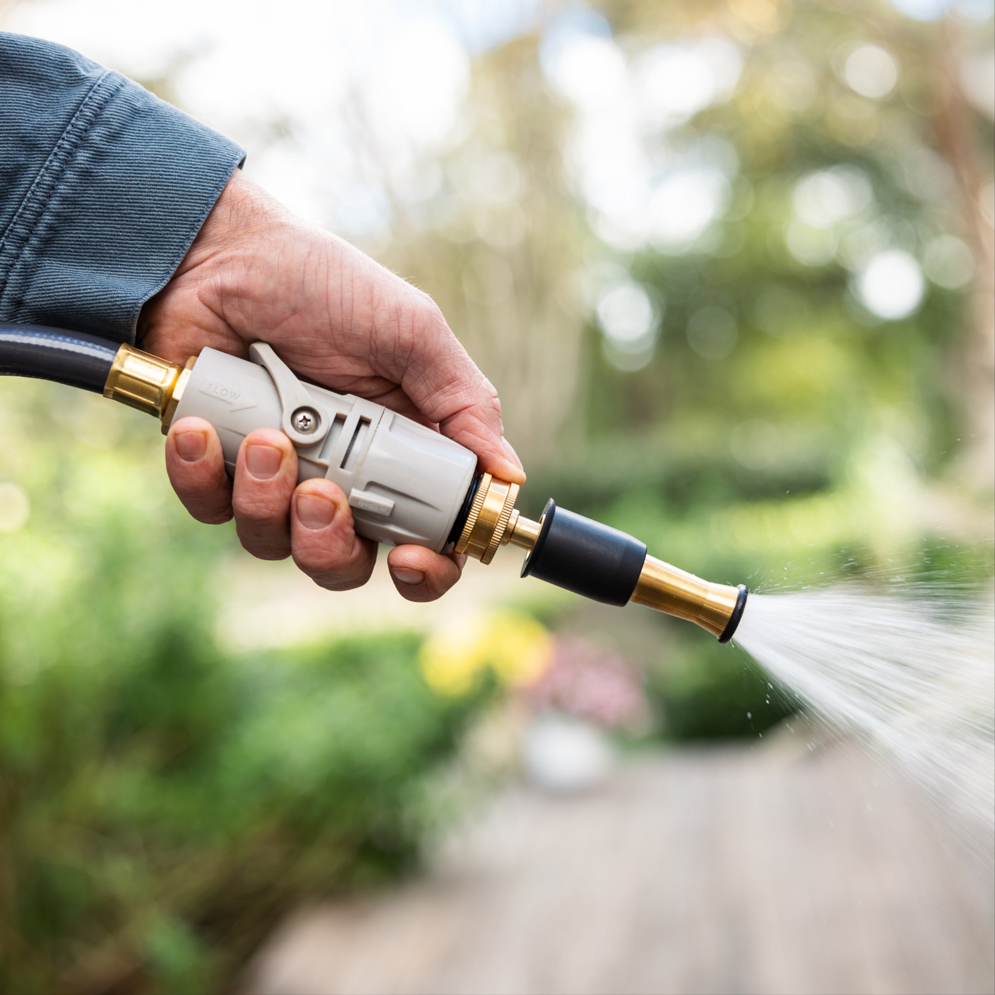 Brass garden hose nozzles held in one hand spraying water with green plants in the background