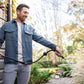 Person wearing blue holding brass garden hose nozzle and spraying a plant with trees in the background